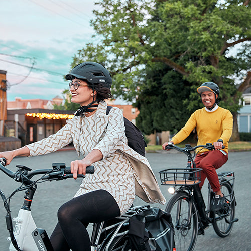 Two smiling people riding electric city bikes down a street 