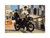 A woman wearing a tan hoodie and black helmet holding the handlebars of a matte black electric bike with a boy wearing a black shirt and jeans and blue helmet sitting on the back. On a bridge in an urban setting.