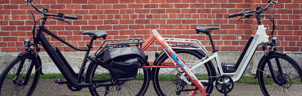 Two ebikes, one black and the other white, against a red brick wall backdrop next to a red bike rack.