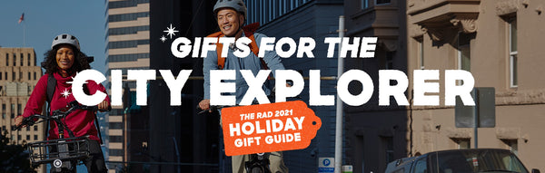 A man and a woman ride up a hill on their electric bikes. A text overlay says "Gifts for the City Explorer" and "the Rad 2021 Holiday Gift Guide"