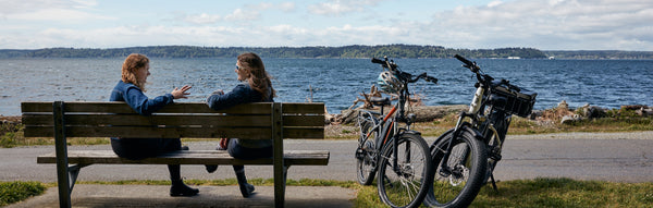 Two women chat on a lakeside park bench next to their electric bikes 