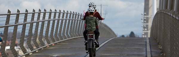 A mother rides a RadRunner Plus on a bridge with her child seated on the back.