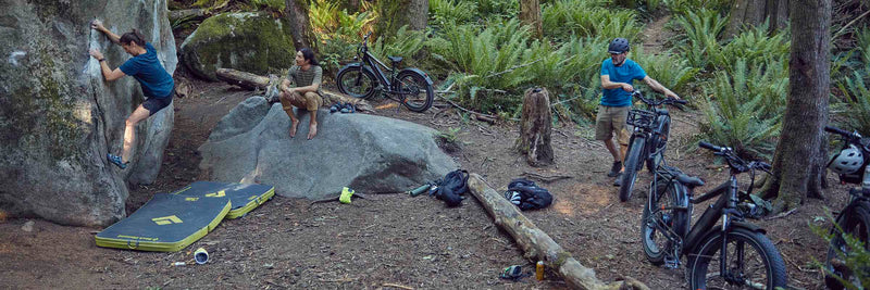 3 people at a rock climbing site in the woods alongside their electric bikes.