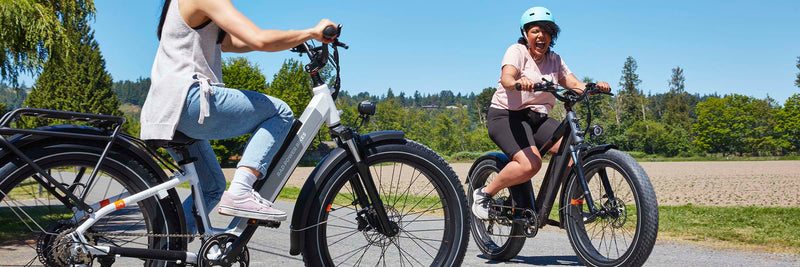 Two women ride their RadRover 6 Plus electric bikes by a farm.