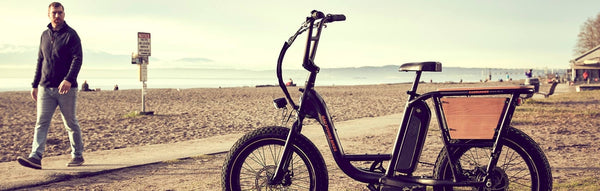 A man on a beach looks over at an electric bike equipped with wood panels.