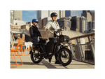 A woman wearing a tan hoodie and black helmet holding the handlebars of a matte black electric bike with a boy wearing a black shirt and jeans and blue helmet sitting on the back. On a bridge in an urban setting. 