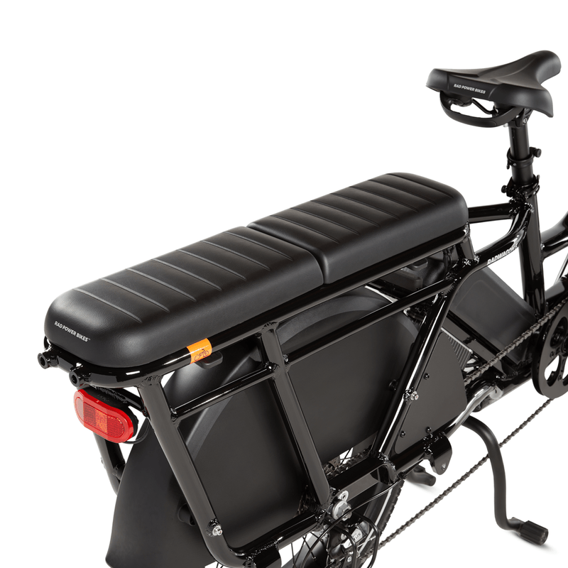 Two RadWagon deckpads installed on the back of a RadWagon electric cargo bike, used for passenger seating. Black vinyl material with padding. Subtle Rad Power Bikes logo along the end.