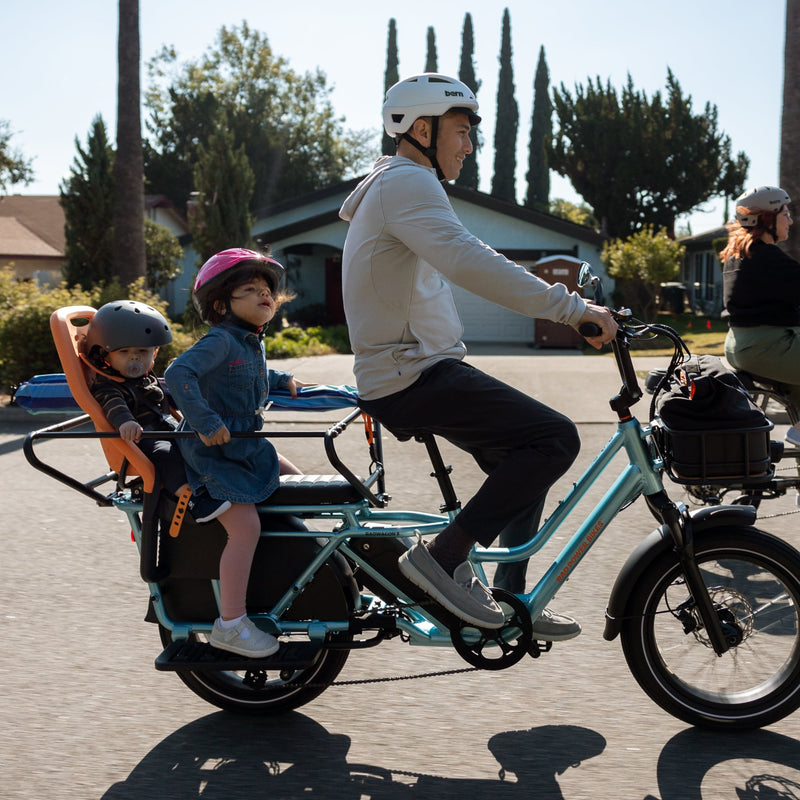 Man riding a blue RadWagon 5 electric cargo bike with two children as passengers. The children are seated with passenger accessories including the RadWagon 5 Deckpad, RadWagon 5 Running Boards, an orange thule yepp child seat and a caboose.