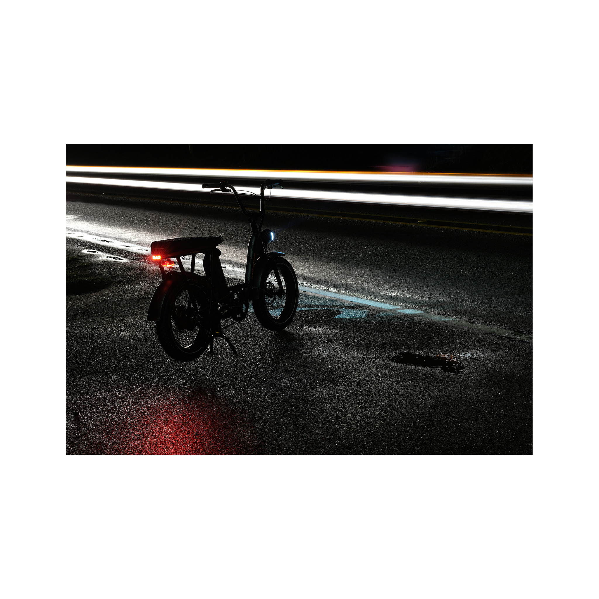 Photo of a Rad Power Bikes RadRunner electric cargo bike with passenger package parked at night on a damp street with Rad logo taillight turned on and mounted below rear seat on the rack.